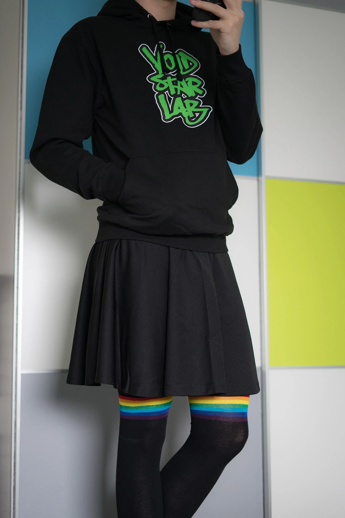 Photo of me standing in front of a wall, wearing a black skirt, a black hoodie with a green graffiti logo on it and black overknee socks with rainbow stripes.