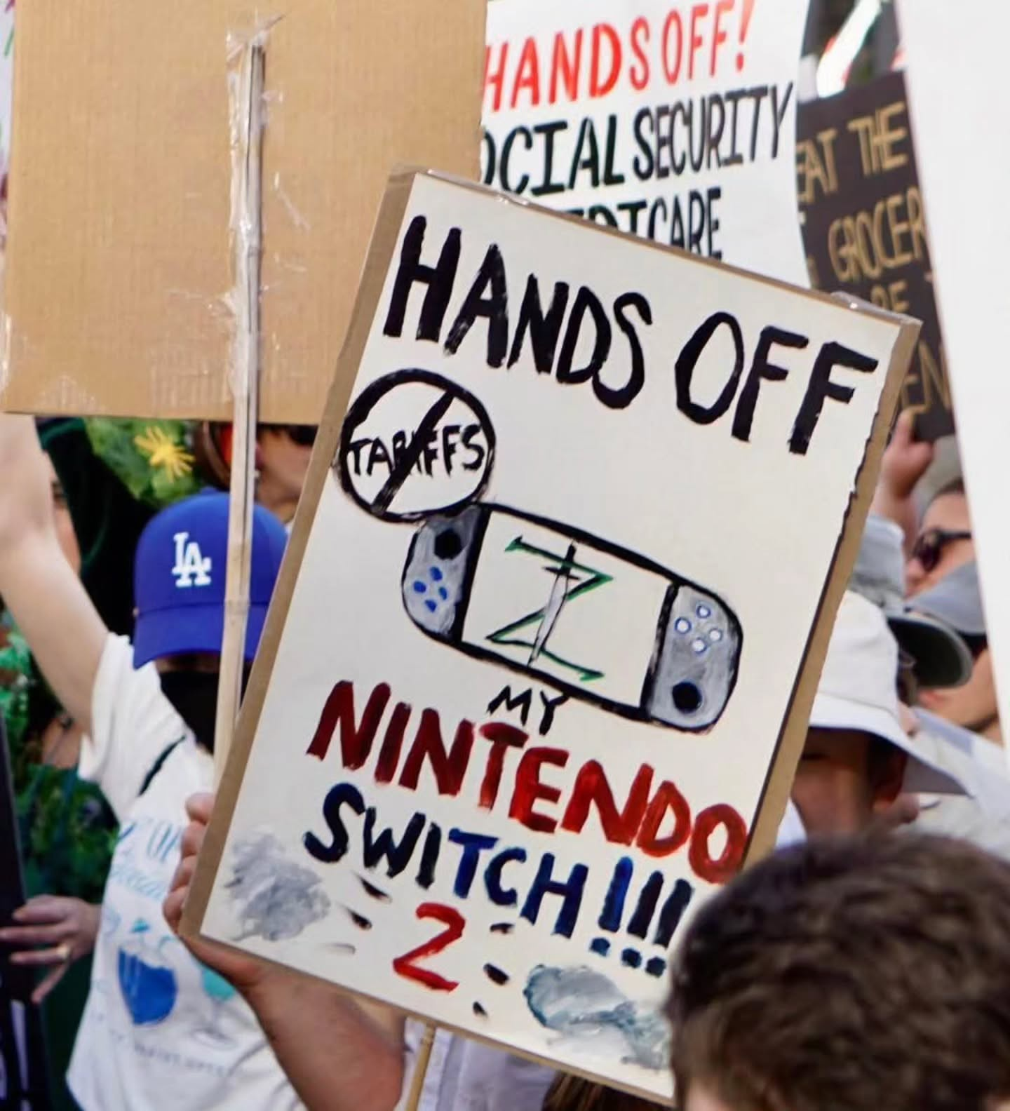 Protester holds a sign with a painted nintendo switch with a Zeld logo on it, an TARRIFS under the international NO sing, and the words HANDS OFF MY NINTENDO SWITCH 2