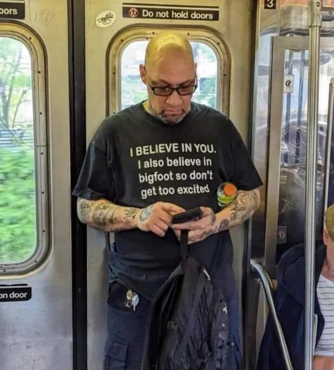 A man on a train, with a black T-shirt that says "I BELIEVE IN YOU. I also believe in bigfoot so don't get too excited"